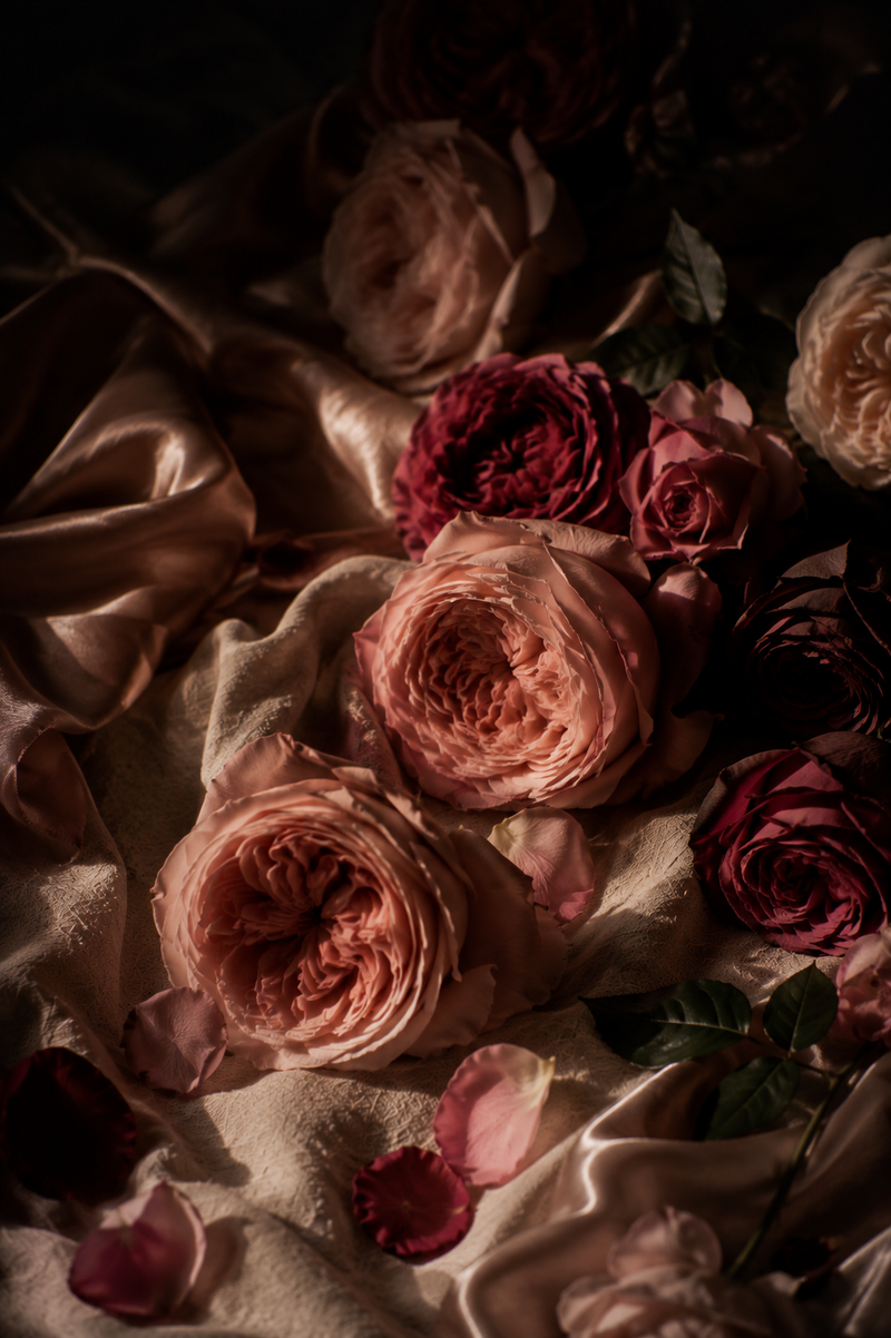 A moody still life arrangement of layered roses in dusty pink, mauve, and deep burgundy tones, draped over cream satin fabric with scattered petals.